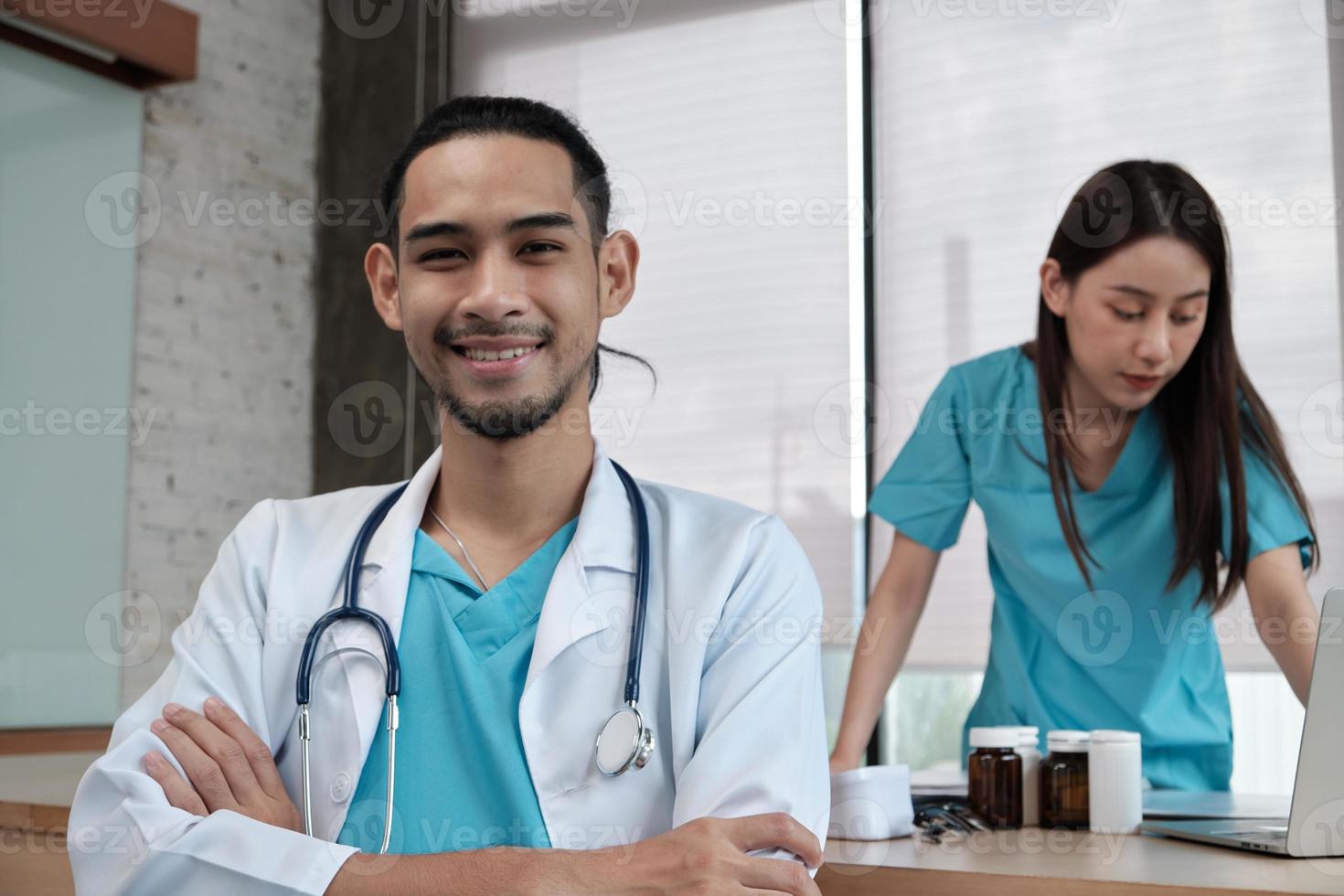 Retrato de joven médico de origen asiático en uniforme con estetoscopio. sonreír y mirar a la cámara en una clínica hospitalaria, pareja femenina trabajando detrás, profesional de medicación para dos personas. foto