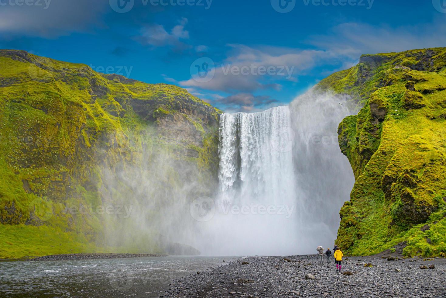 poderosa y famosa cascada de skogafoss con una persona de pie solitaria con chaqueta naranja, mientras camina en islandia, verano, vista escénica dramática en un día soleado y cielo azul. foto