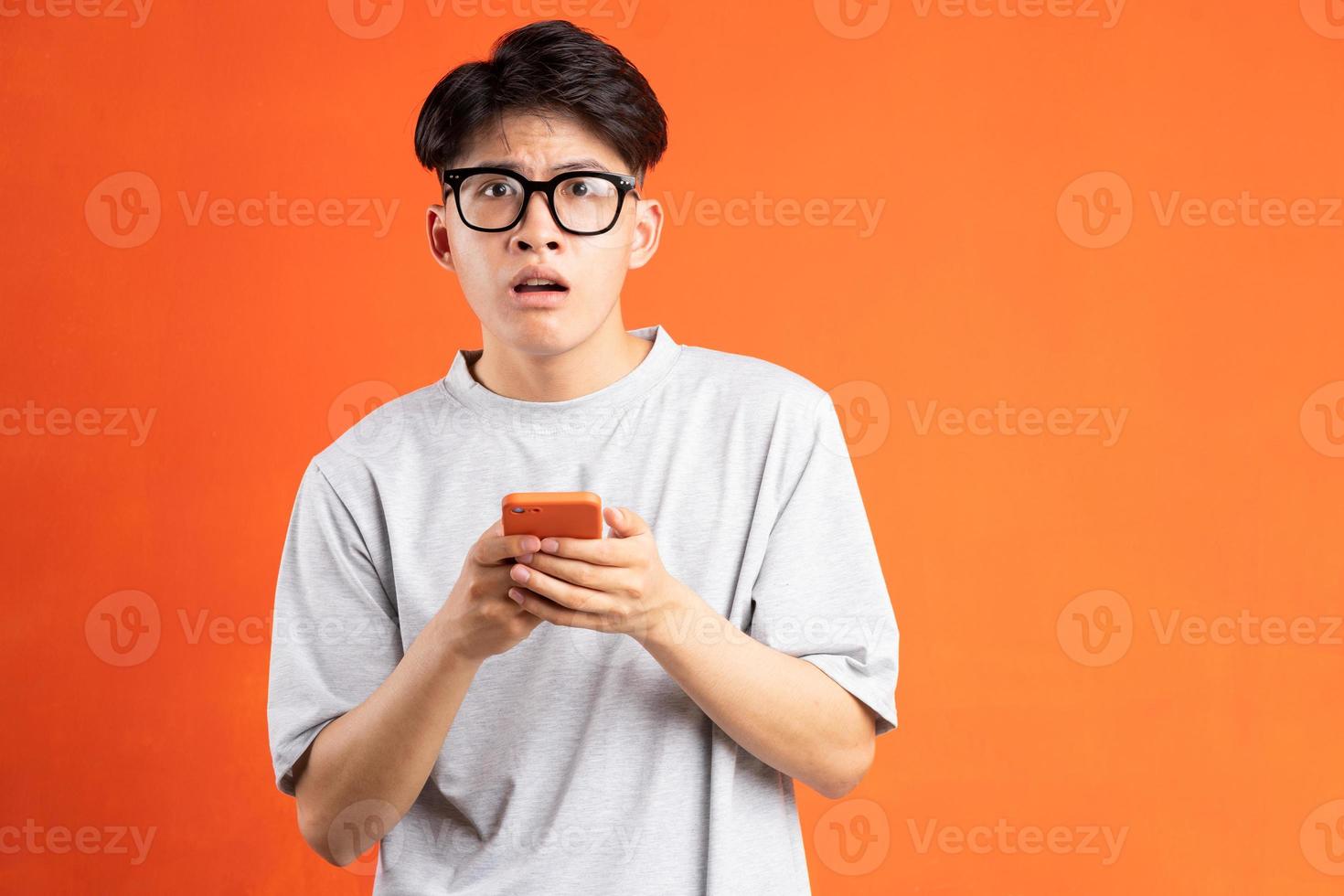 Portrait of young asian man holding smartphone in hand with cryptic expression, isolated on orange background photo