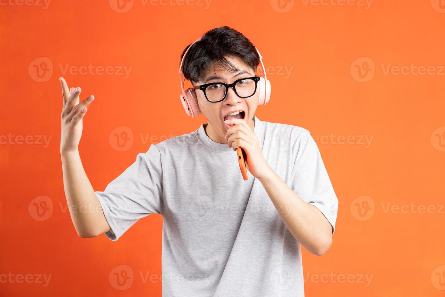 Portrait of young asian man holding smart phone in hand wearing headphones and singing, isolated on orange background photo