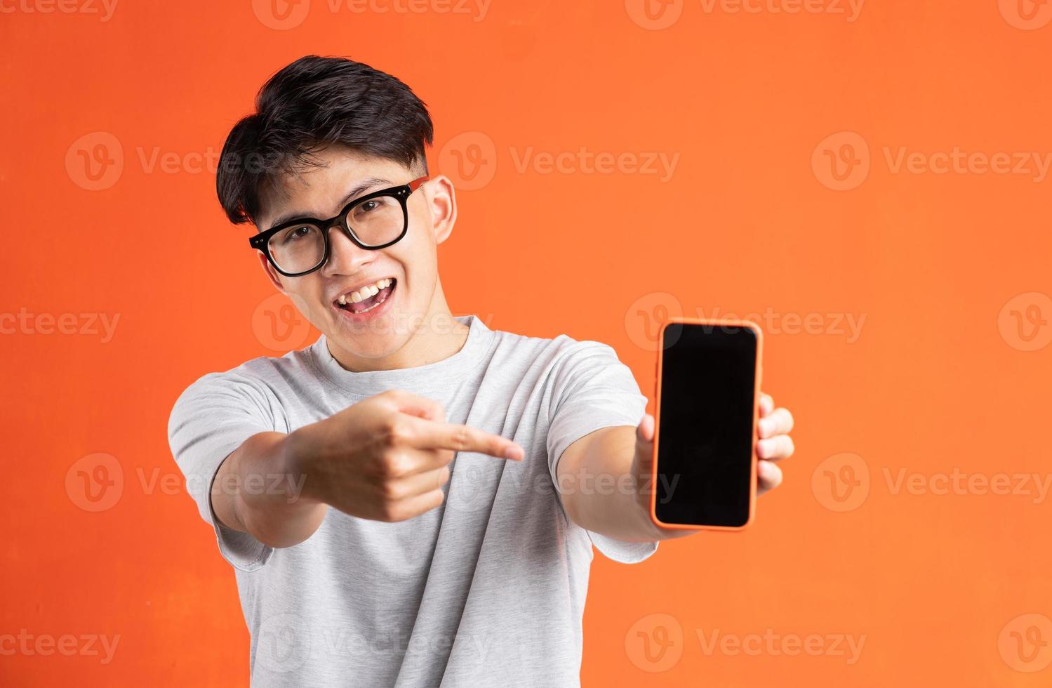 Portrait of young asian man pointing at phone screen, isolated on orange background photo