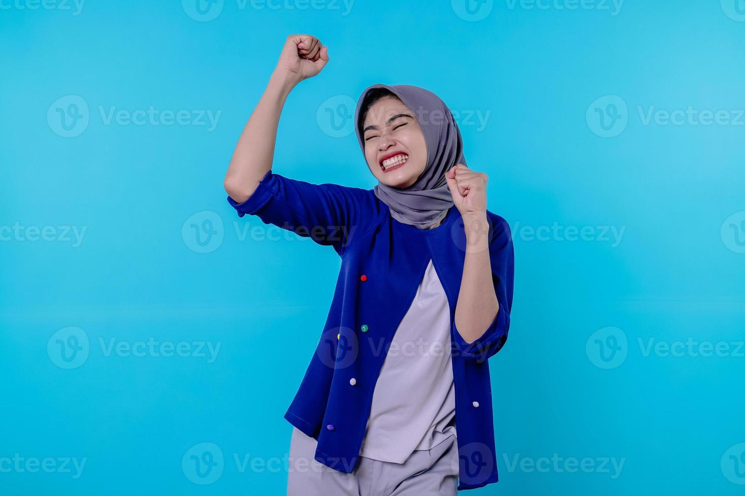 Cheerful young female has positive expression, clenches fists, has overjoyed look, being in high spirit, wearing hijab, isolated over blue wall photo