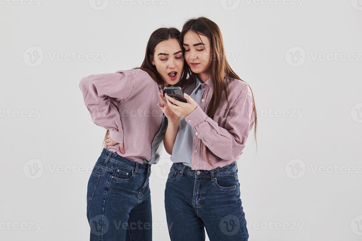 Some interesting content in their phones. Sharing with secrets. Two sisters twins standing and posing in the studio with white background photo