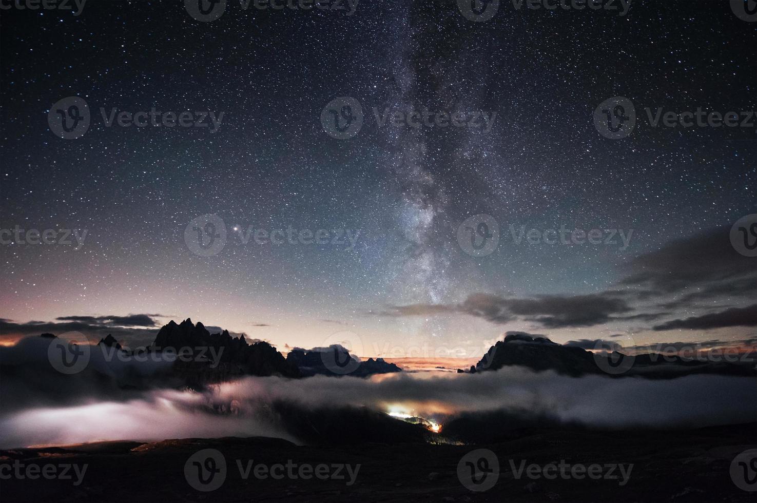 You can clear see the milky way. Something is shining in the middle of the forest with stars in the sky. The mountains are surrounded by dense fog photo