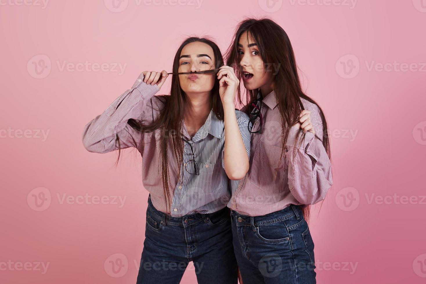 haciendo bigote con pelo. mujeres jóvenes divirtiéndose en el estudio con fondo rosa. gemelos adorables foto