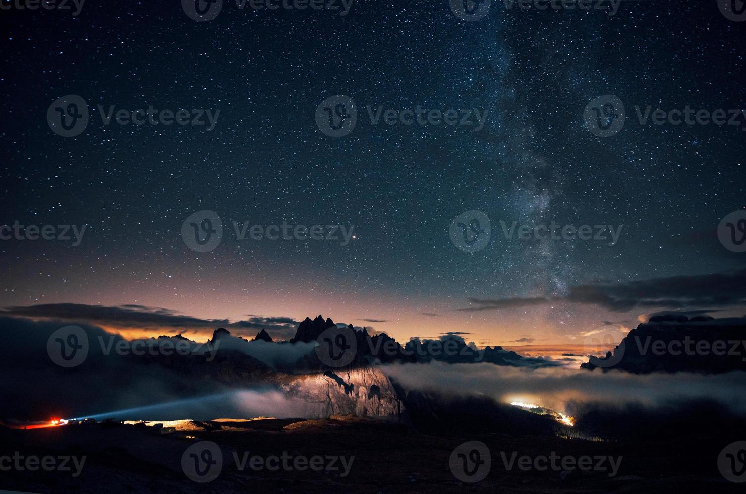 luces de vehículos de larga distancia que vienen en el lado izquierdo. hermoso espacio lleno de estrellas en el cielo. las montañas están rodeadas de una densa niebla foto