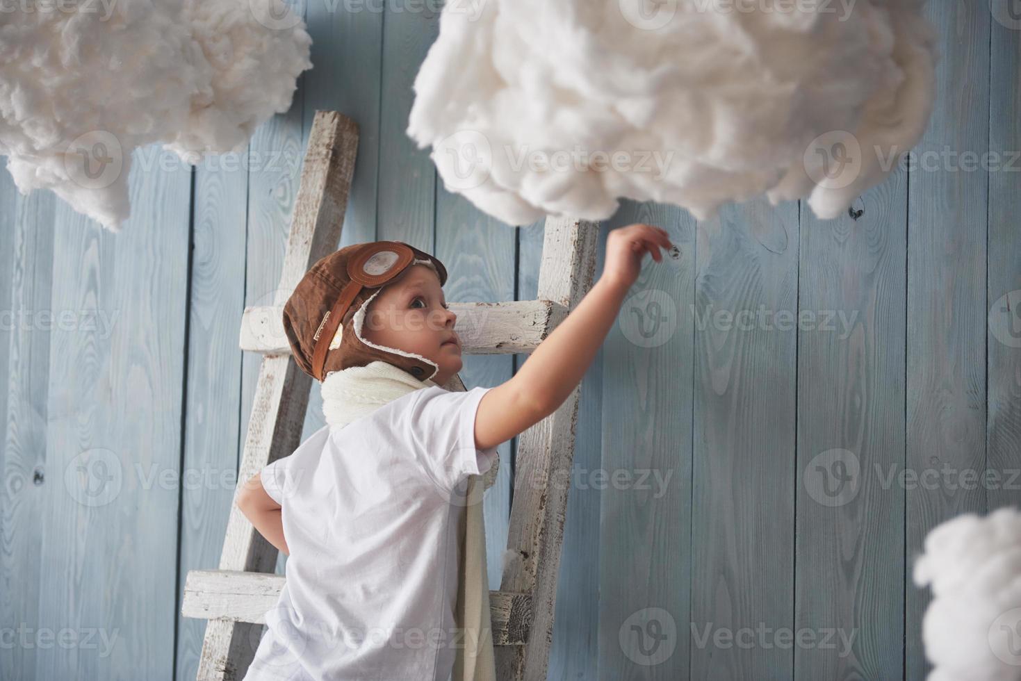 niño con sombrero de piloto de pie en la escalera en el estudio. llegar al cielo. tocar el concepto de nubes foto