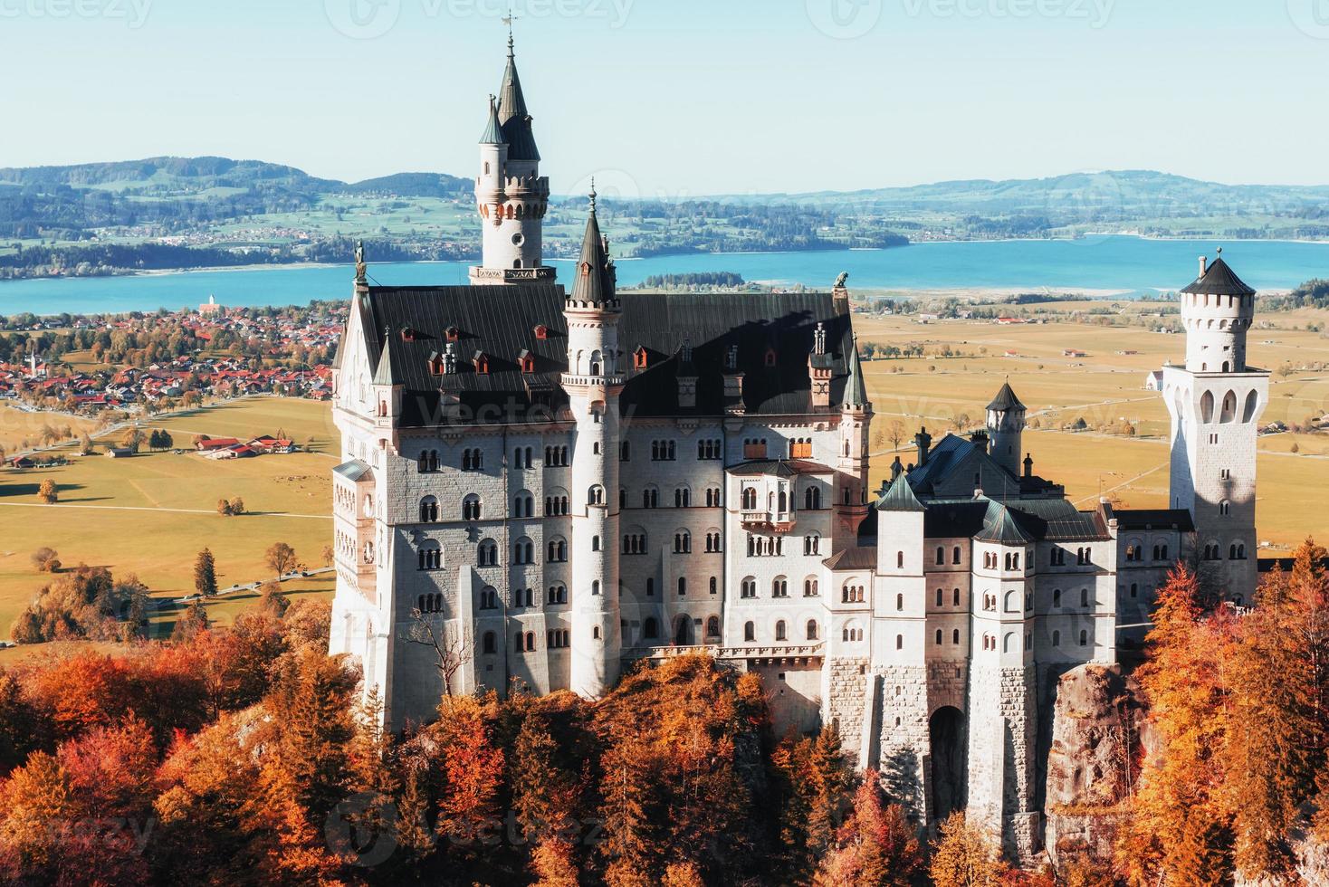 Gran castillo de Neuschwanstein excepcional de pie en la llanura con árboles debajo y una pequeña ciudad en el fondo foto