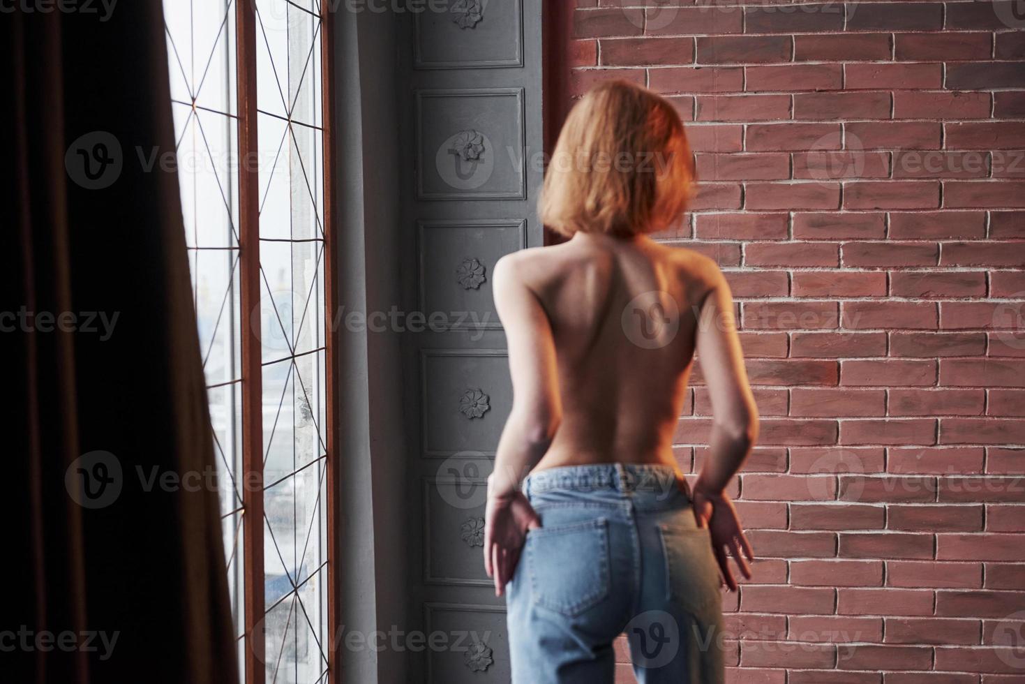 Rear view. Hot young blonde with bare chest and jeans stands against the window and brick wall photo