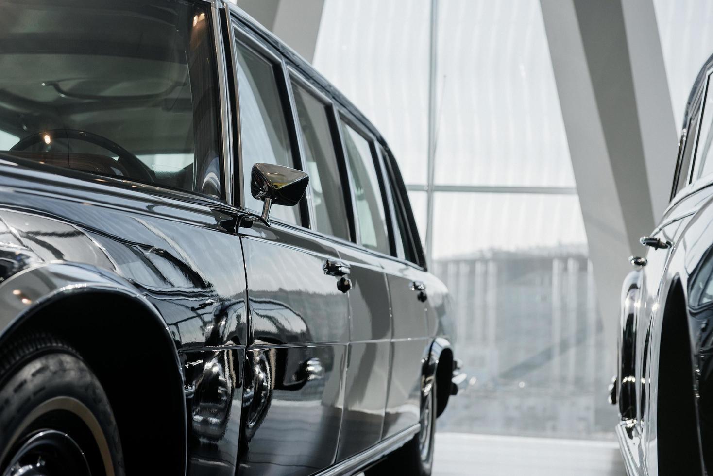 View of driver's side black retro business class limousine with chrome mirror parked near another vintage car in a light building photo