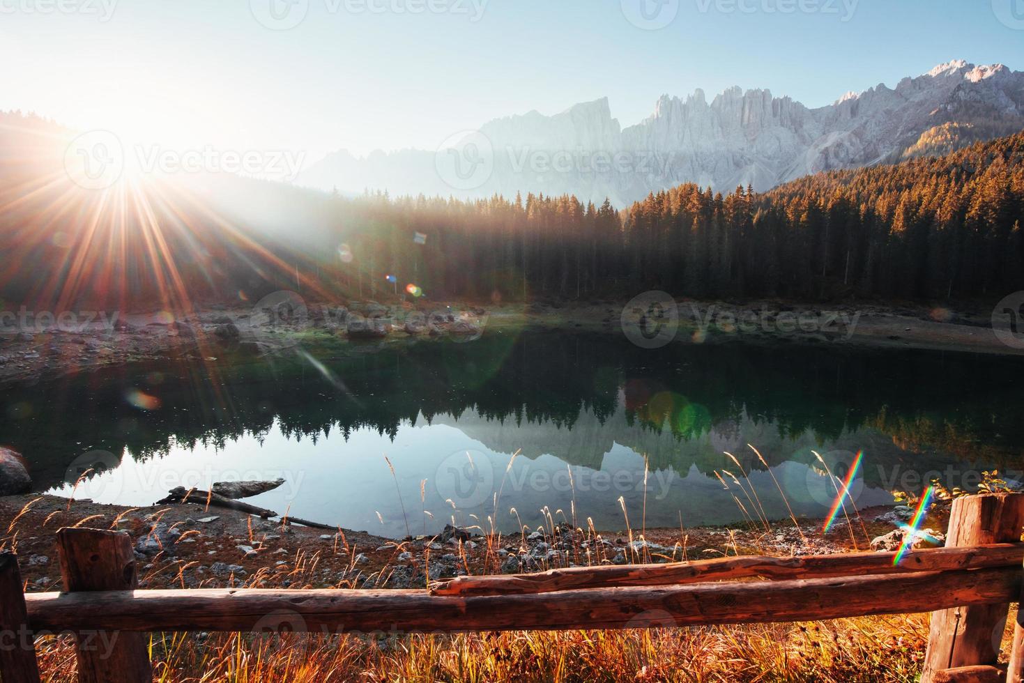 Straight light beam into the camera. Wooden fence. Autumn landscape with clear lake, fir forest and majestic mountains photo