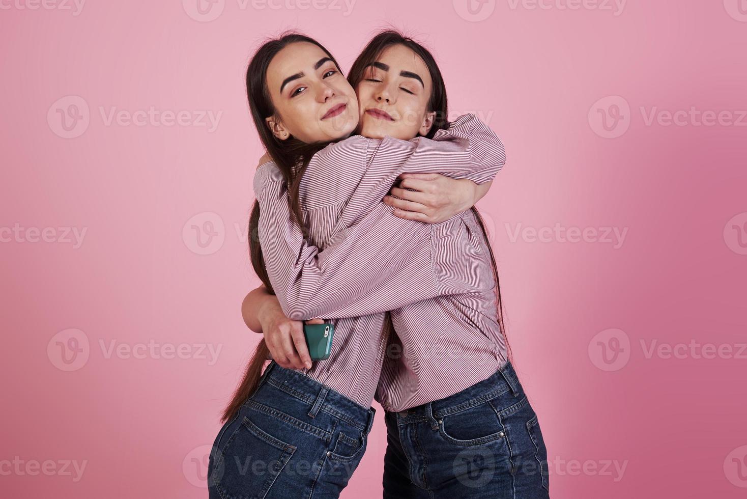 Abrazos familiares entre dos hermanas en ropa idéntica en el estudio con fondo de color rosa foto