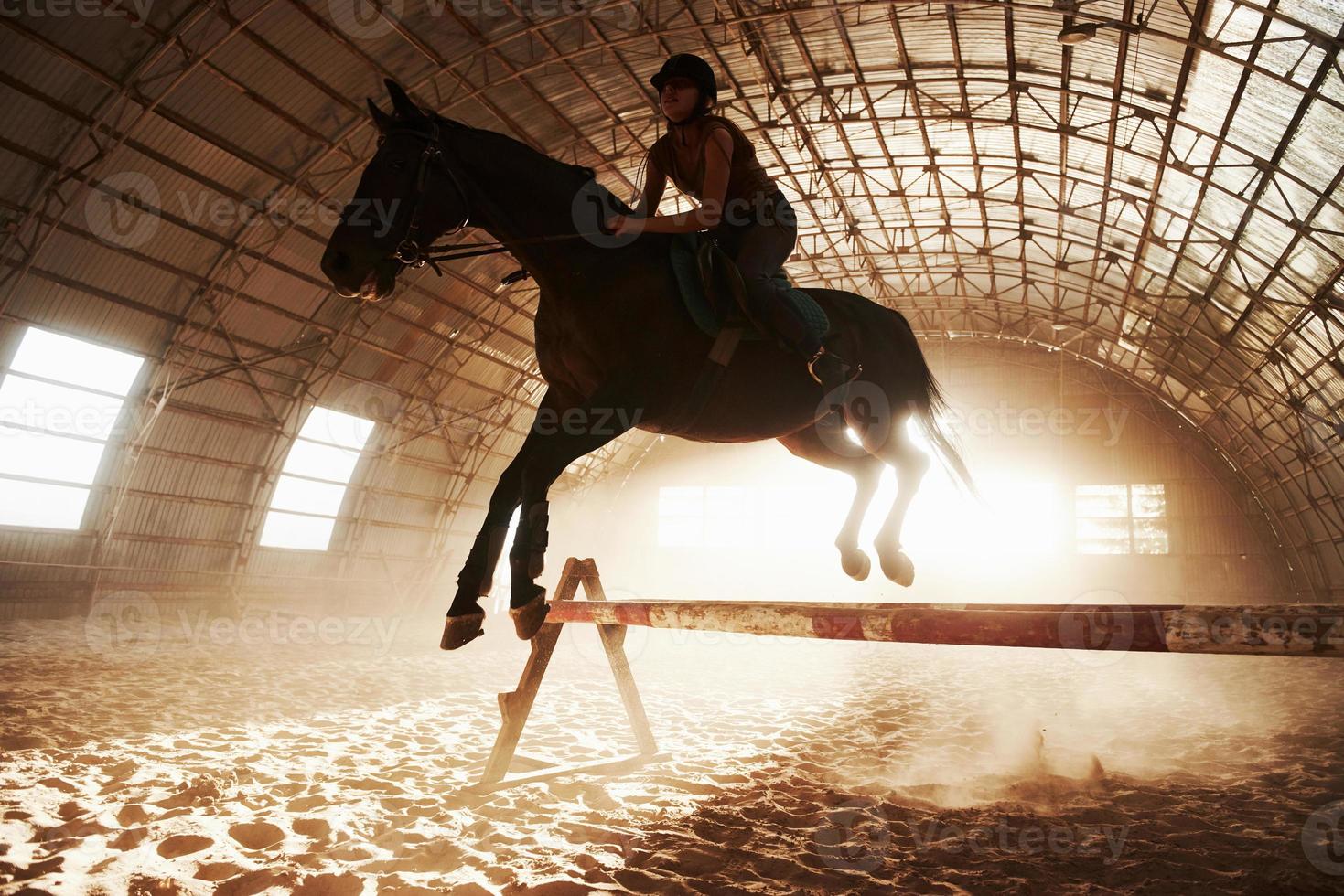 majestuosa imagen de caballo caballo silueta con jinete sobre fondo puesta de sol. la chica jockey a lomos de un semental cabalga en un hangar en una granja y salta por encima del travesaño. el concepto de montar a caballo foto