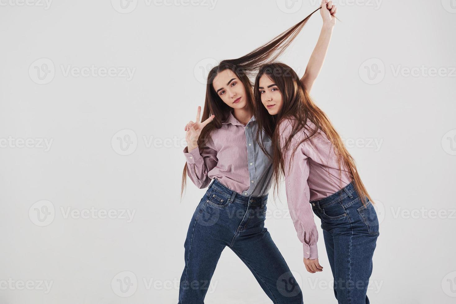 Left girl showing gesture of two fingers. Two sisters twins standing and posing in the studio with white background photo