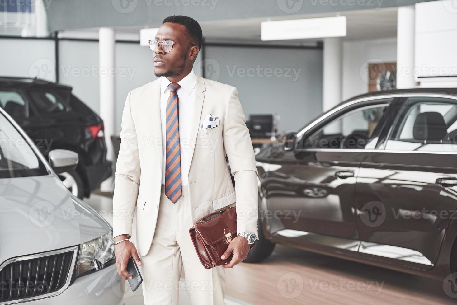 joven empresario negro en el fondo del salón del automóvil. concepto de venta y alquiler de coches foto