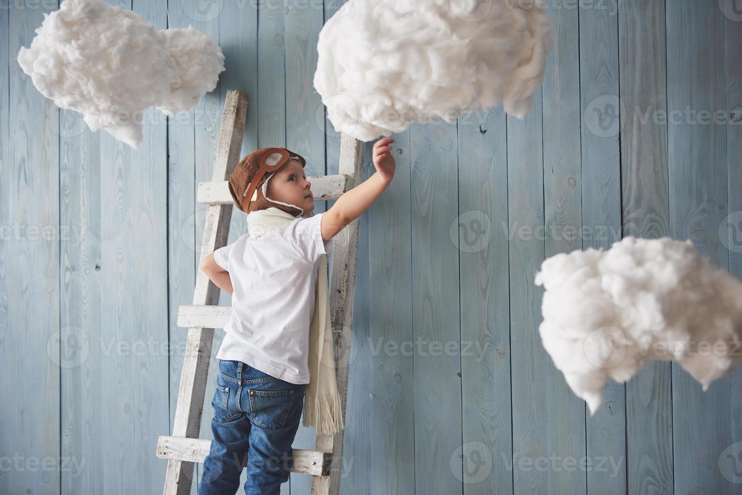 Little boy in pilot hat standing on the ladder in the studio. Reach to heaven. Touch the clouds concept photo