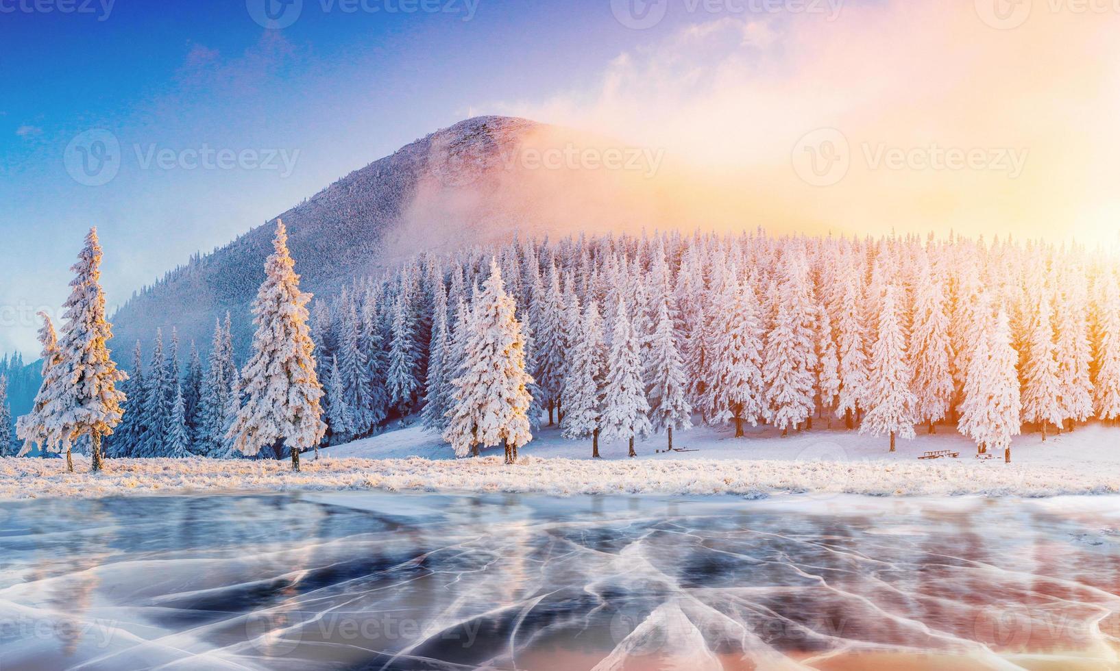 Cracks on the surface of the blue ice. Frozen lake in winter mountains. It is snowing. The hills of pines. Carpathian Ukraine Europe photo