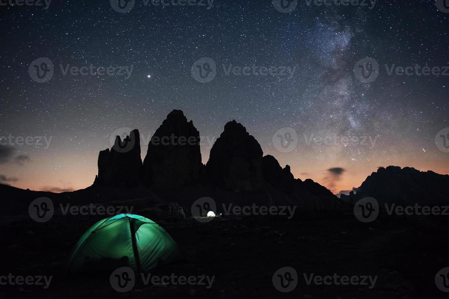 nuestra galaxia está en el cielo. Iluminación de dos carpas con turistas en el interior cerca de las montañas tre cime tres picos durante la noche foto