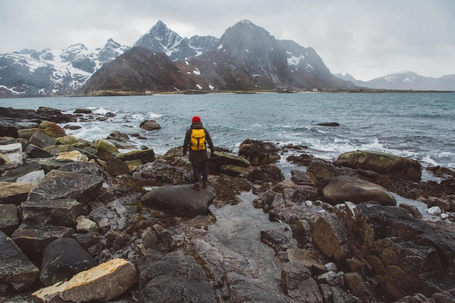 Traveler man with a yellow backpack wearing a red hat standing on the background of mountains and sea. Travel lifestyle concept. Shoot from the back photo