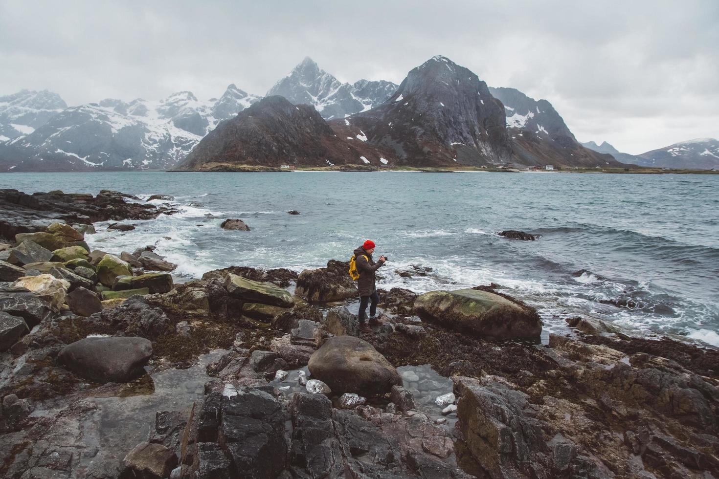 Traveller professional photographer taking nature photo of landscape. Wearing a yellow backpack wearing a red hat standing on rocks against the background of the sea and the mountains