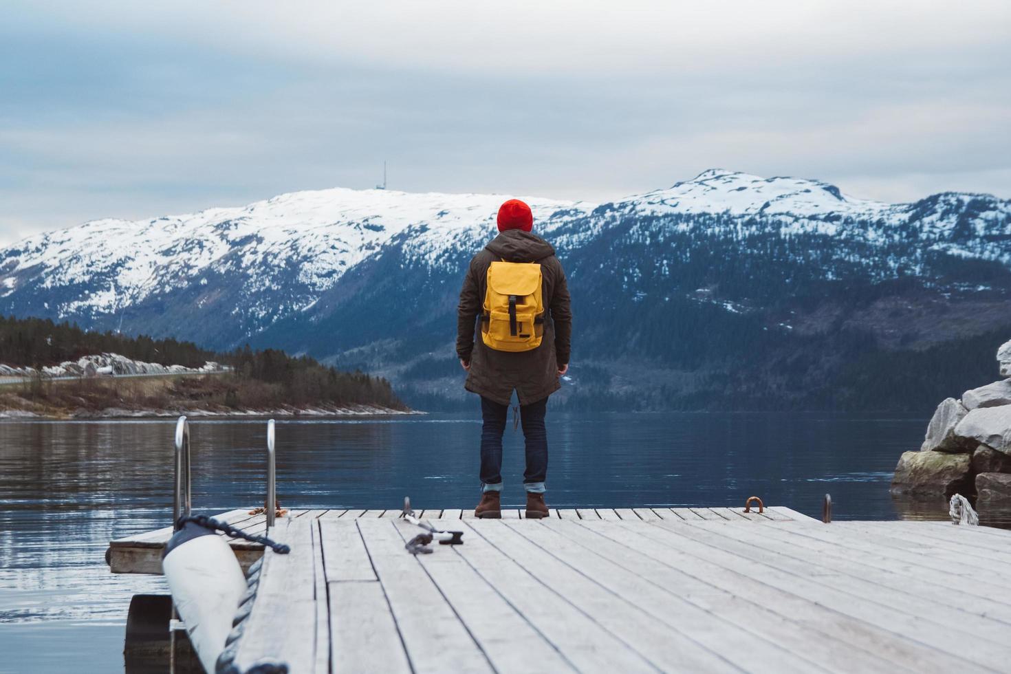 Traveler man with a yellow backpack wearing a red hat standing on the background of mountain and lake wooden pier. Travel lifestyle concept. Shoot from the back photo