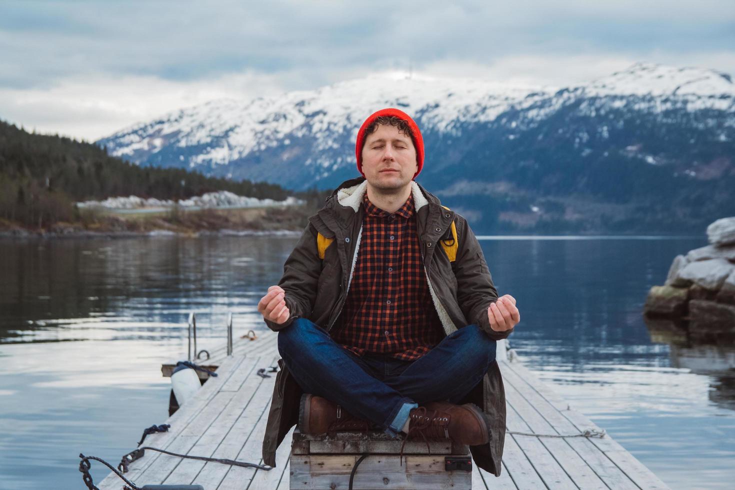 Traveler man in a meditative position sitting on a wooden pier on the background of a mountain and a lake. Space for your text message or promotional content. photo