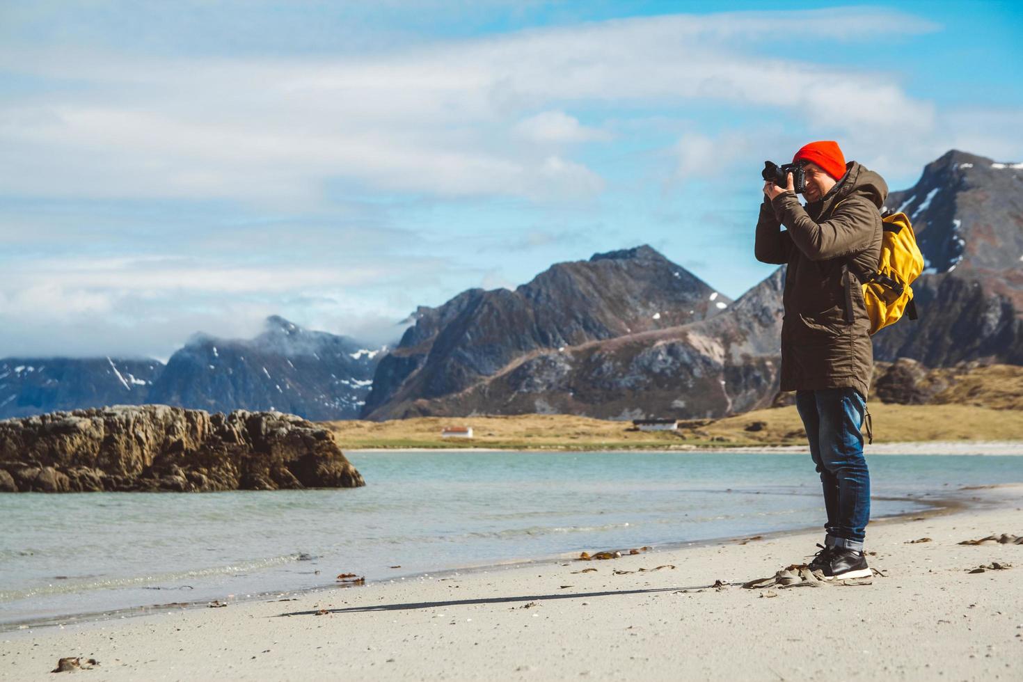 Traveler is a professional photographer taking over the landscape photo landscape. Wearing a yellow backpack in a red hat standing on a sandy beach on the background of the sea and mountains