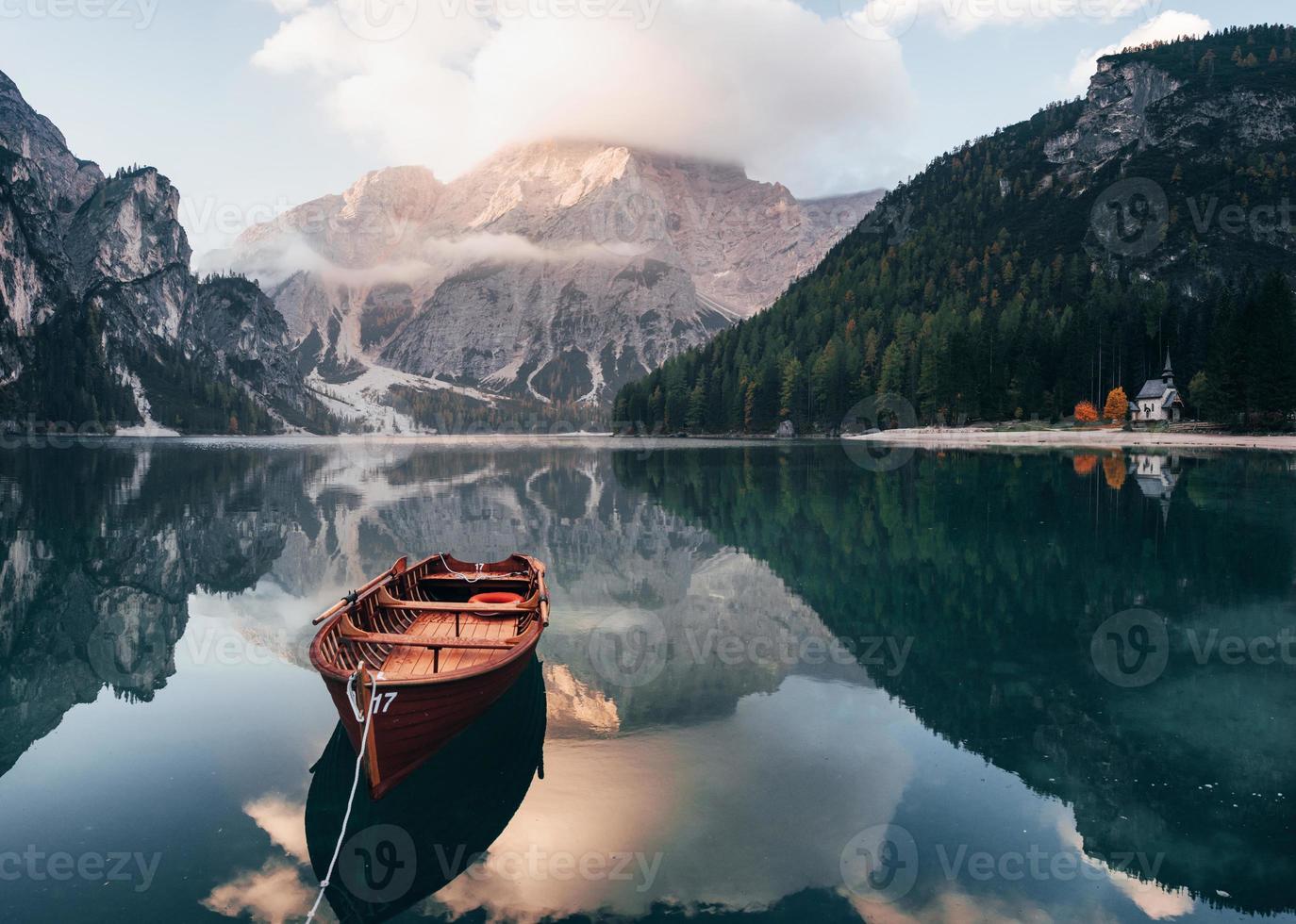 su luz del sol en la cima. barco de madera en el lago de cristal con majestuosa montaña detrás. reflejo en el agua. la capilla está en la costa derecha foto