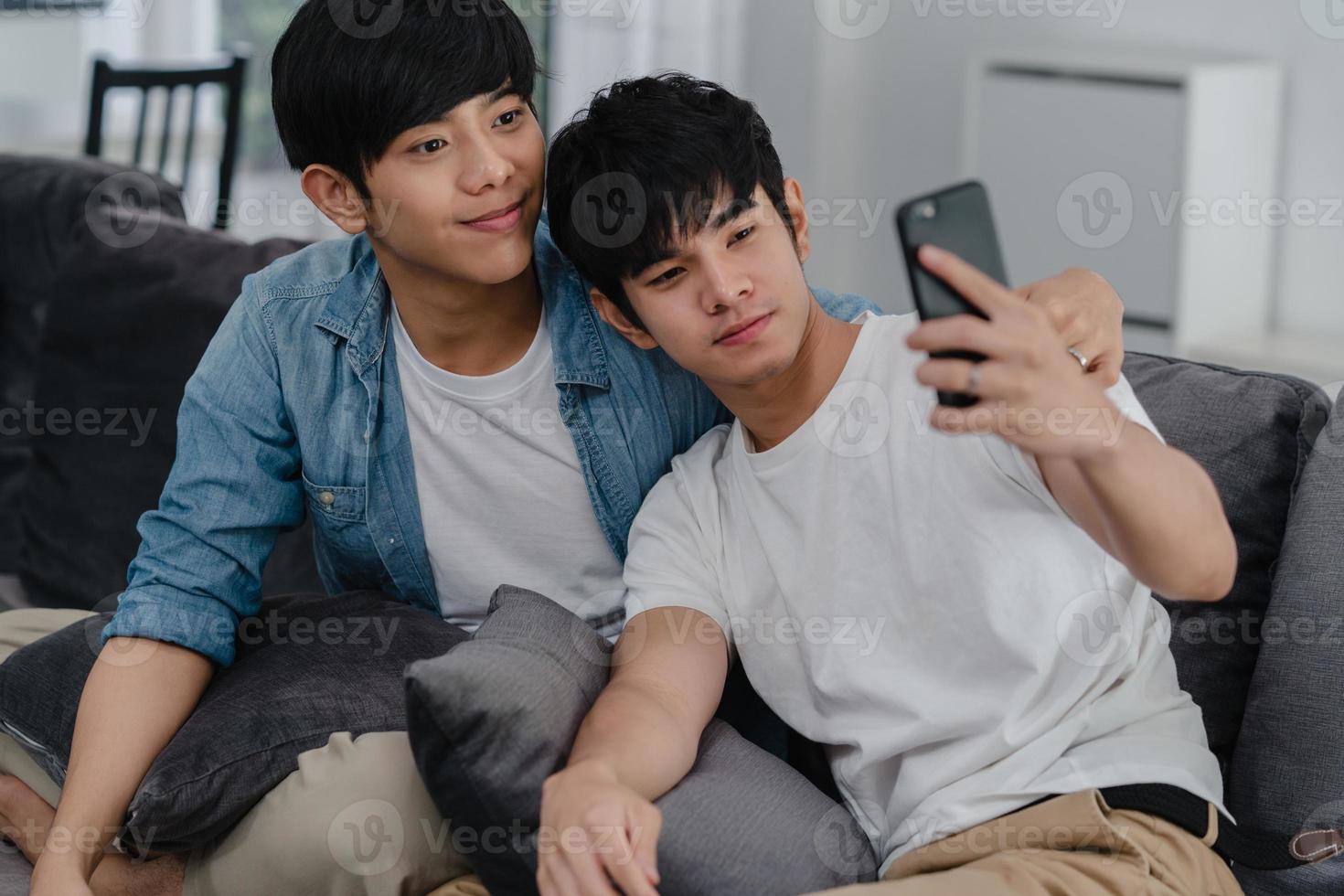 Asian influencer Gay couple vlog at home. Asian LGBTQ men happy relax fun using technology mobile phone record lifestyle vlog video upload in social media while lying sofa in living room concept. photo
