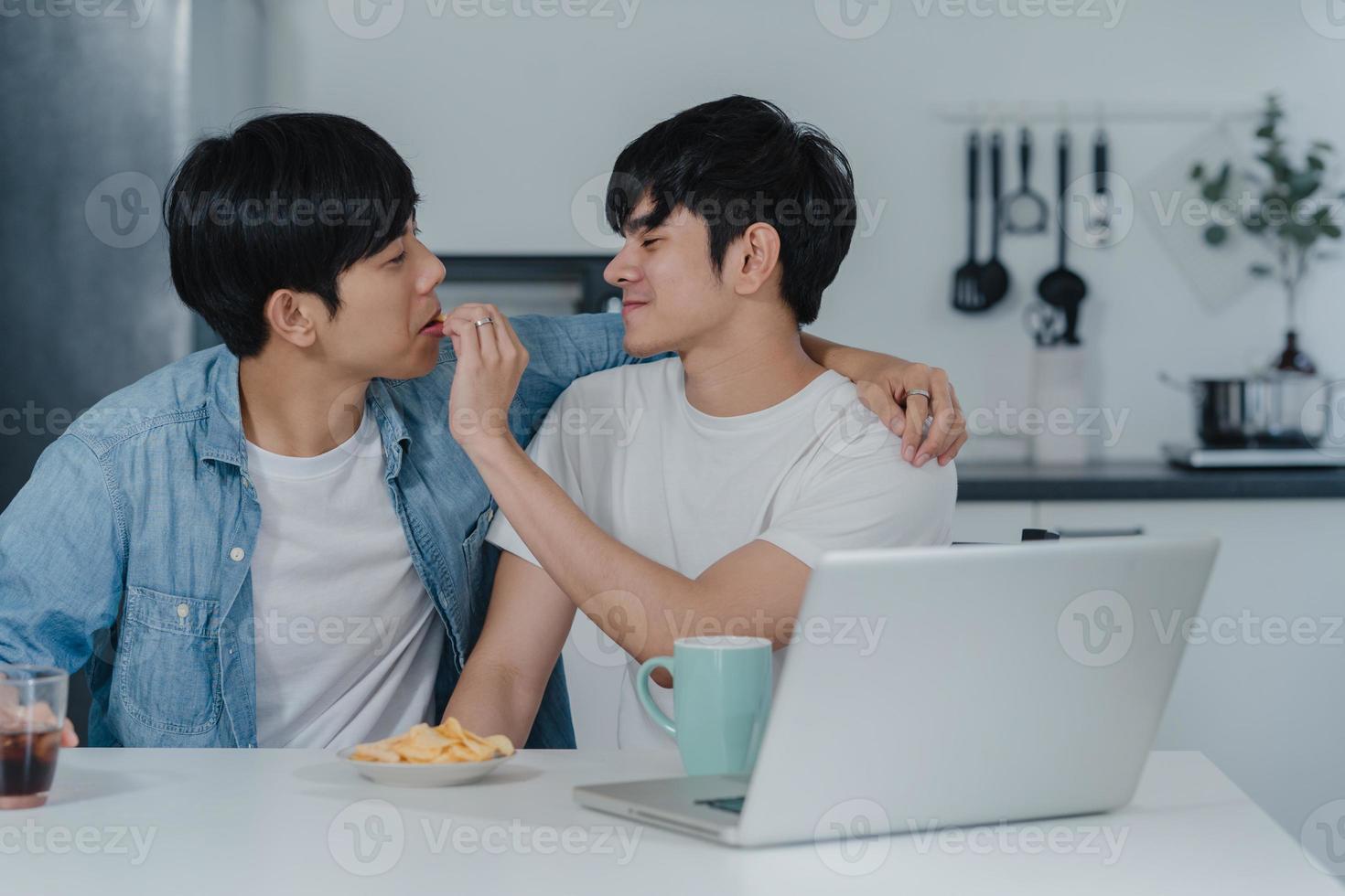 Young Gay couple feeding food and snack using computer laptop at modern home. Asian LGBTQ men happy relax fun using technology social media together while sitting table in kitchen at house concept. photo