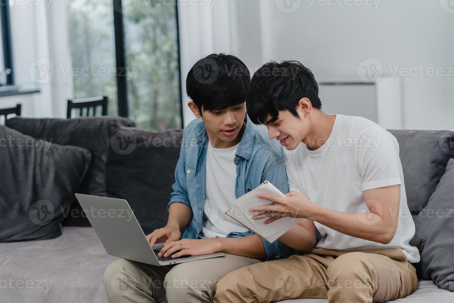 Young Asian Gay couple working laptop at modern home. Asia LGBTQ men happy relax fun using computer and analyzing their finances in internet together while lying sofa in living room at house concept. photo