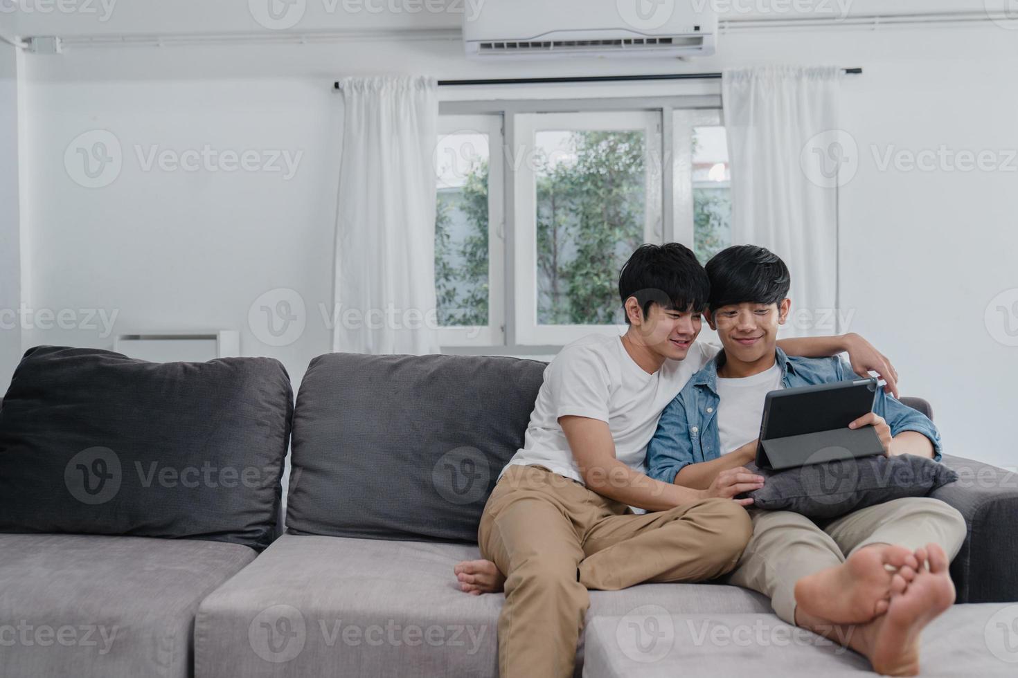 joven pareja gay con tableta en casa. los hombres asiáticos lgbtq felices se relajan y se divierten usando la tecnología viendo películas en Internet juntos mientras están acostados en el sofá en el concepto de sala de estar. foto