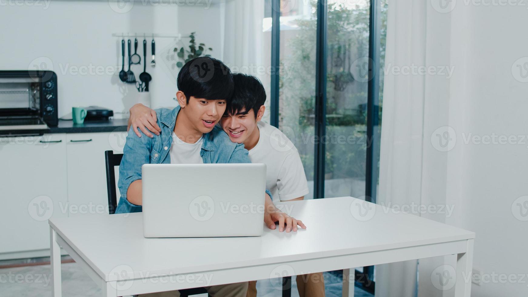 Young Gay couple using computer laptop at modern home. Asian LGBTQ men happy relax fun using technology watching movie in internet together while sitting table in kitchen at house concept. photo