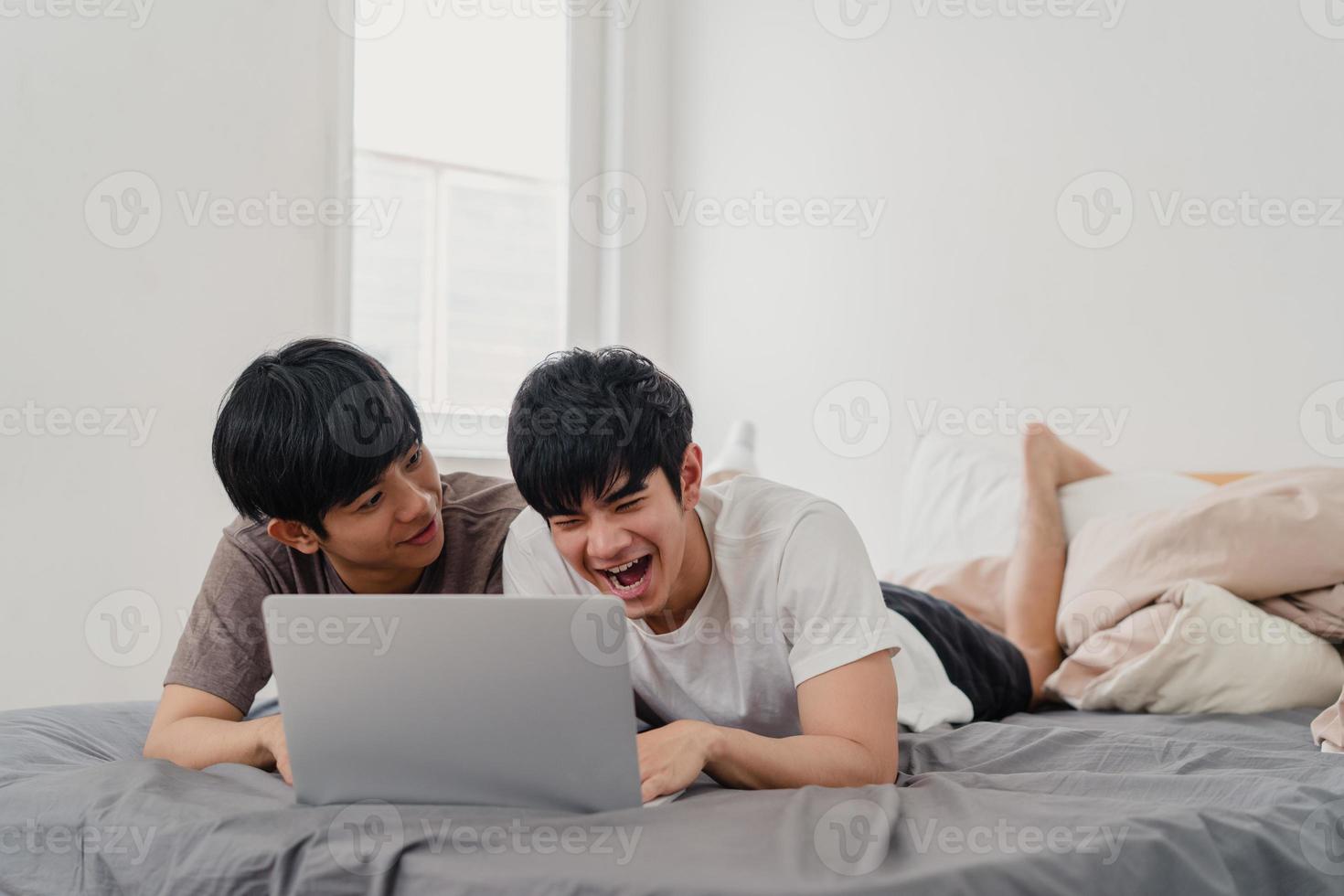 Asian Gay lgbtq men couple using computer laptop at modern home. Young Asia lover male happy relax rest together after wake up, watching movie lying on bed in bedroom at house in the morning concept. photo