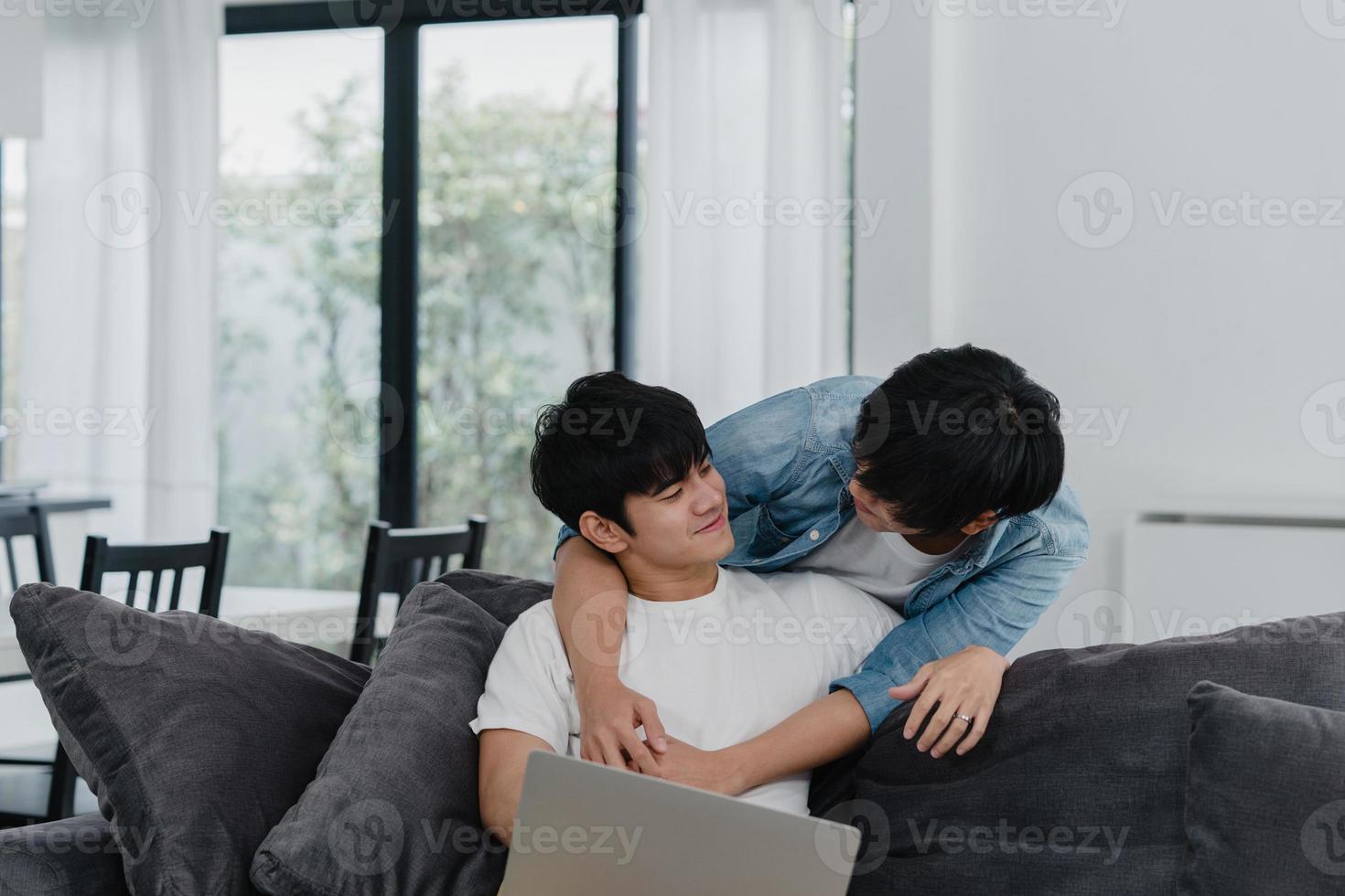 Young Gay couple using computer laptop at modern home. Asian LGBTQ men happy relax fun using technology watching movie in internet together while lying sofa in living room at house concept. photo