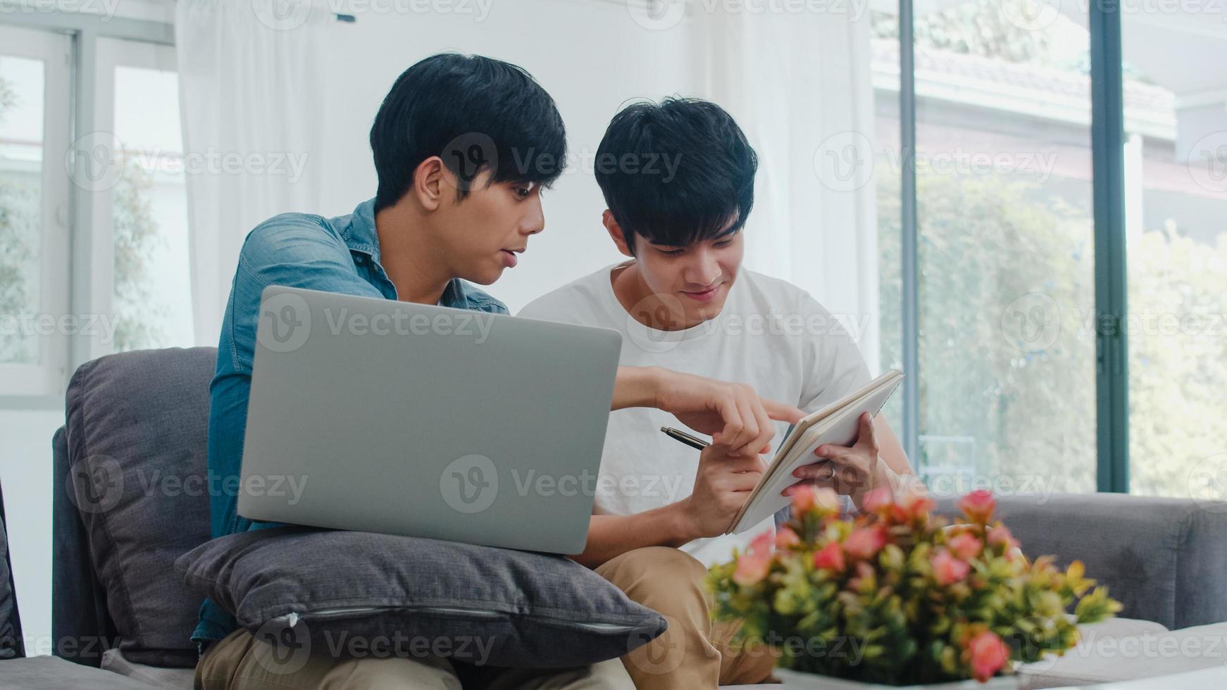 Young Asian Gay couple working laptop at modern home. Asia LGBTQ men happy relax fun using computer and analyzing their finances in internet together while lying sofa in living room at house concept. photo