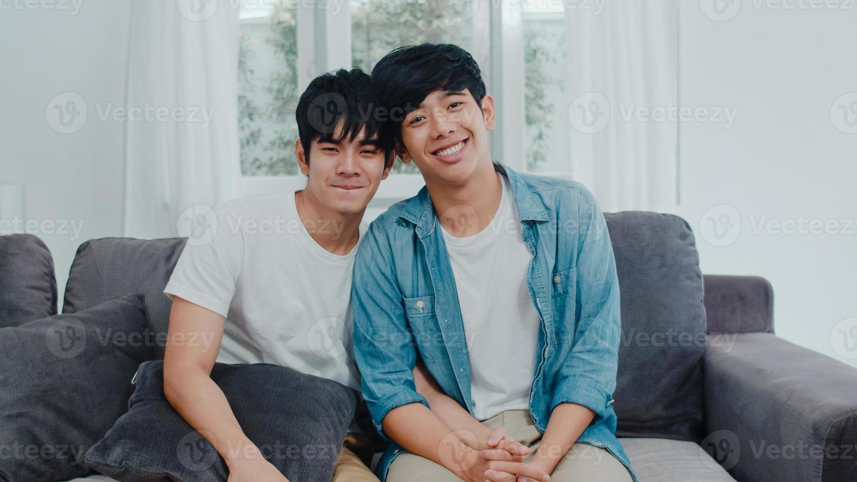 retrato joven pareja gay asiática sentirse feliz sonriendo en casa. los hombres asiáticos lgbtq relajan una sonrisa con dientes mirando a la cámara mientras están acostados en el sofá en la sala de estar en casa en el concepto de la mañana. foto