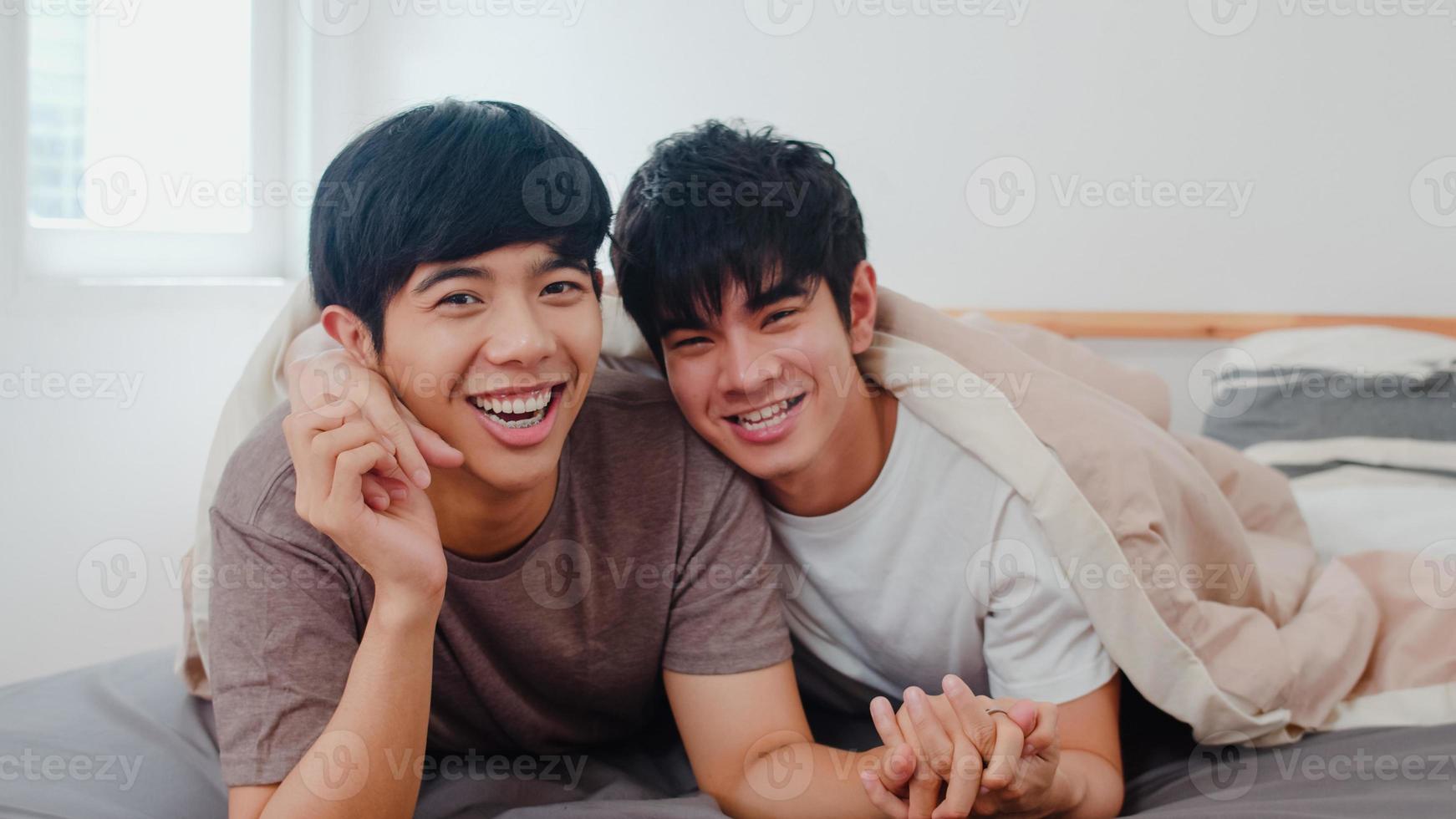 retrato joven pareja gay asiática que se siente feliz en casa. Los hombres lgbtq de Asia se relajan con una sonrisa con dientes mirando a la cámara mientras descansan juntos y pasan un momento romántico después de despertarse en el dormitorio de la casa moderna por la mañana. foto