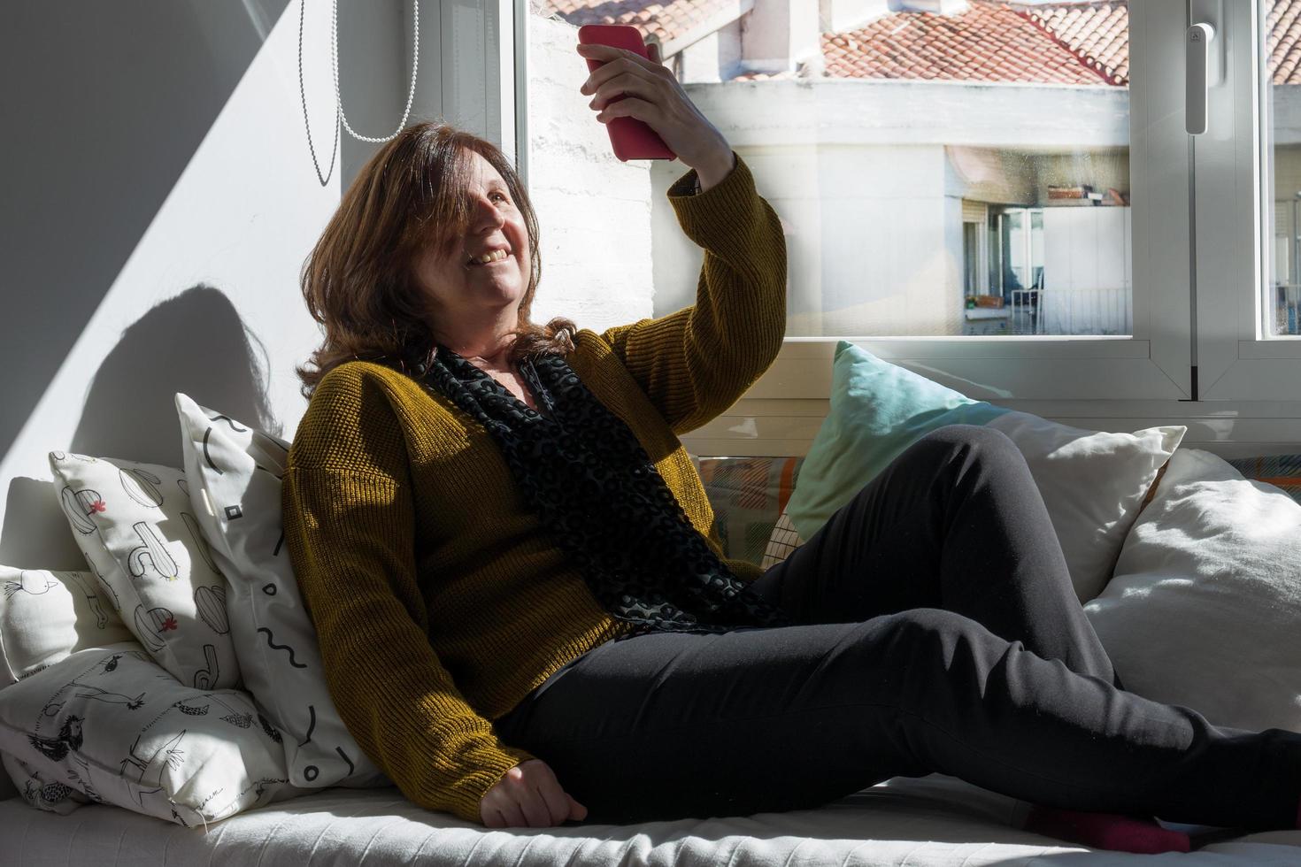 mujer caucásica de 50 años sonriendo durante un video chat con un teléfono. jersey amarillo y pantalón negro. en un sofá cerca de la ventana. foto