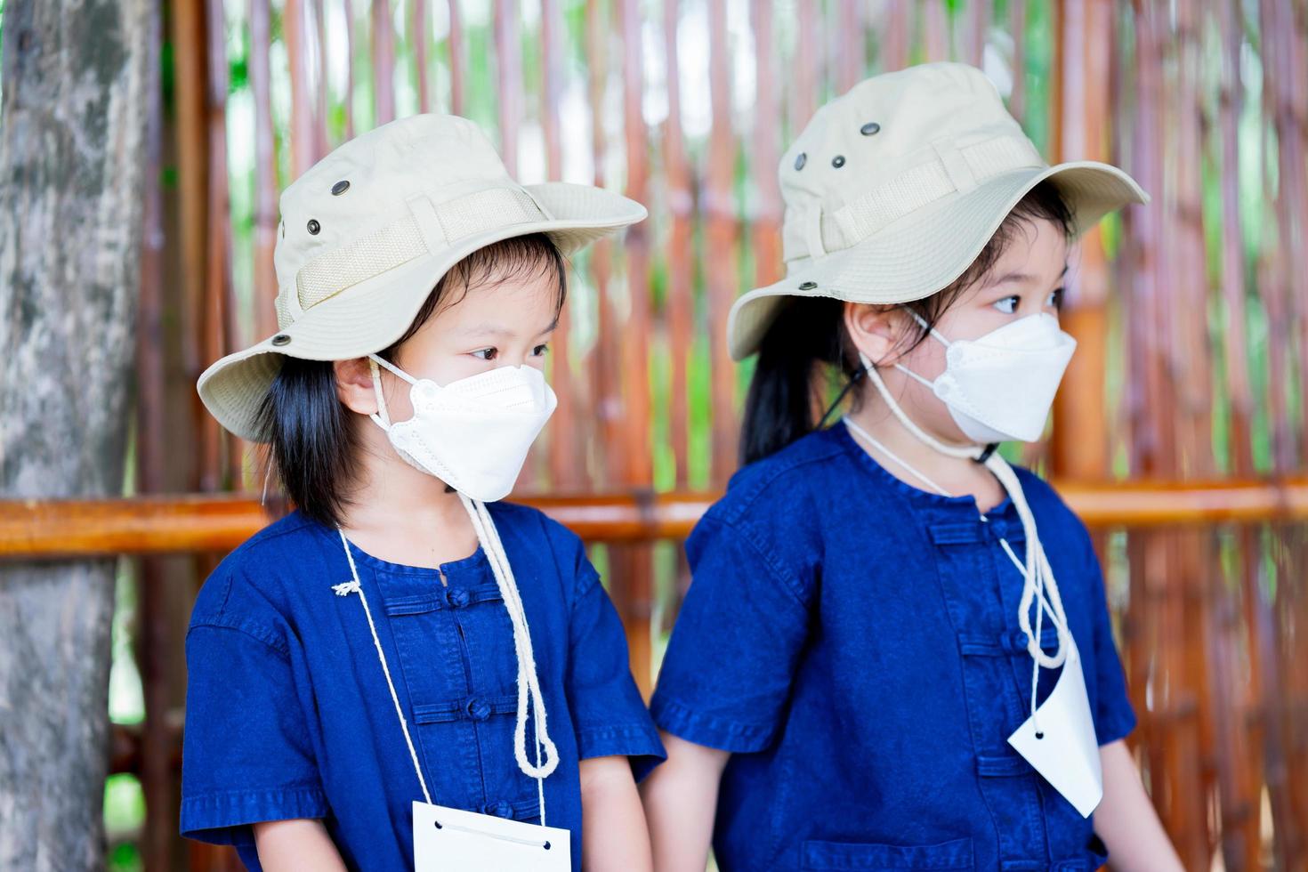 Kindergarten kid wearing 4D white surgical face mask, cream hat and blue Mohamed suit. Pupil sitting on bamboo amphitheater. Learning outside the classroom concept. photo