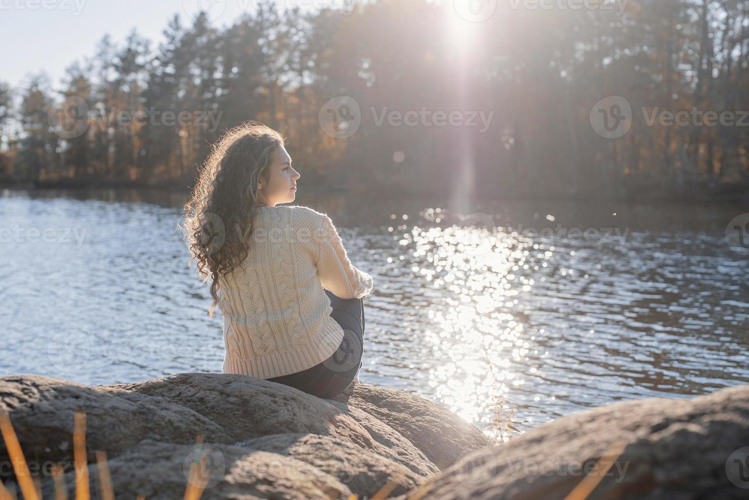 Mujer romántica pensativa sentada en la orilla del río al atardecer en día de otoño foto