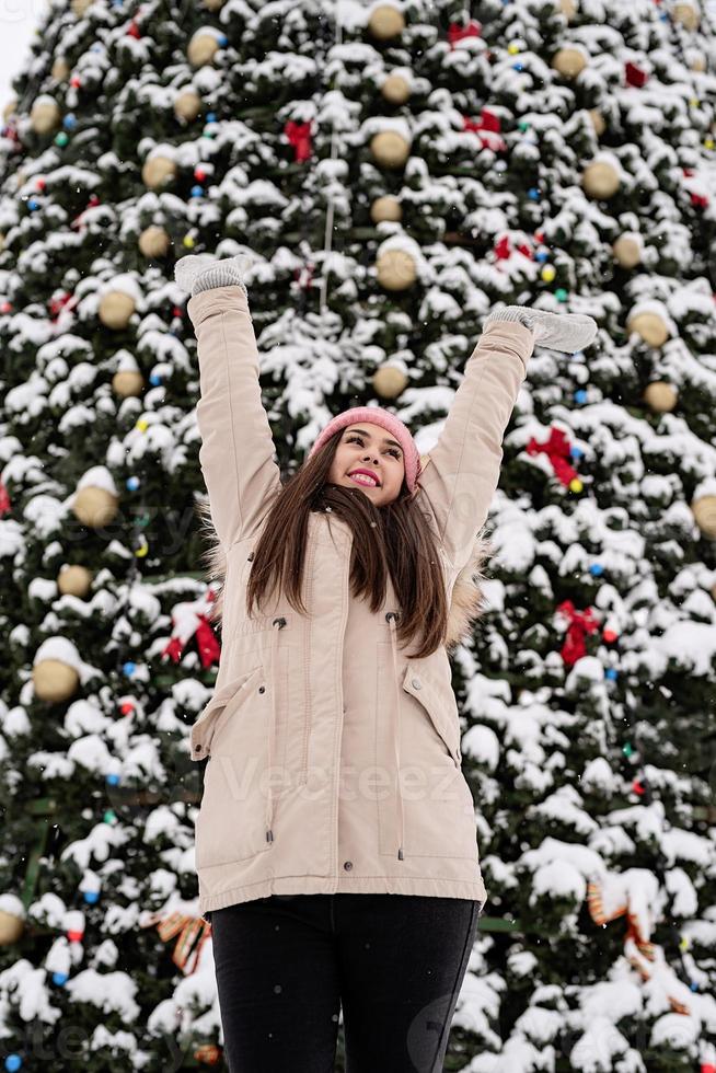 Woman in warm winter clothes standing by the big christmas tree outdoors, arms up, snow falling photo