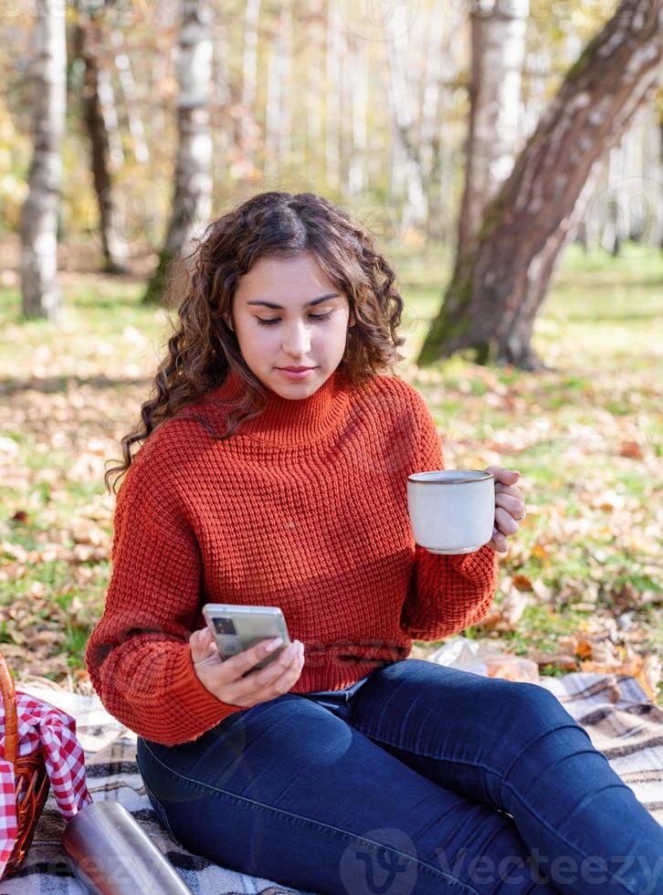 Beautiful woman in red sweater on a picnic in a autumn forest using mobile photo