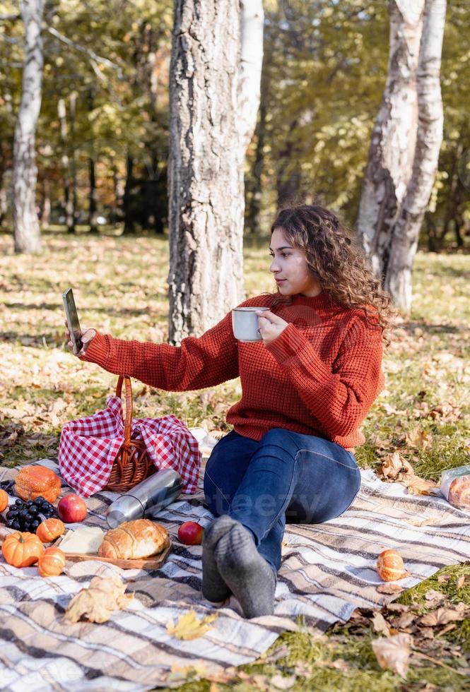 Beautiful woman in red sweater on a picnic in a autumn forest using mobile photo