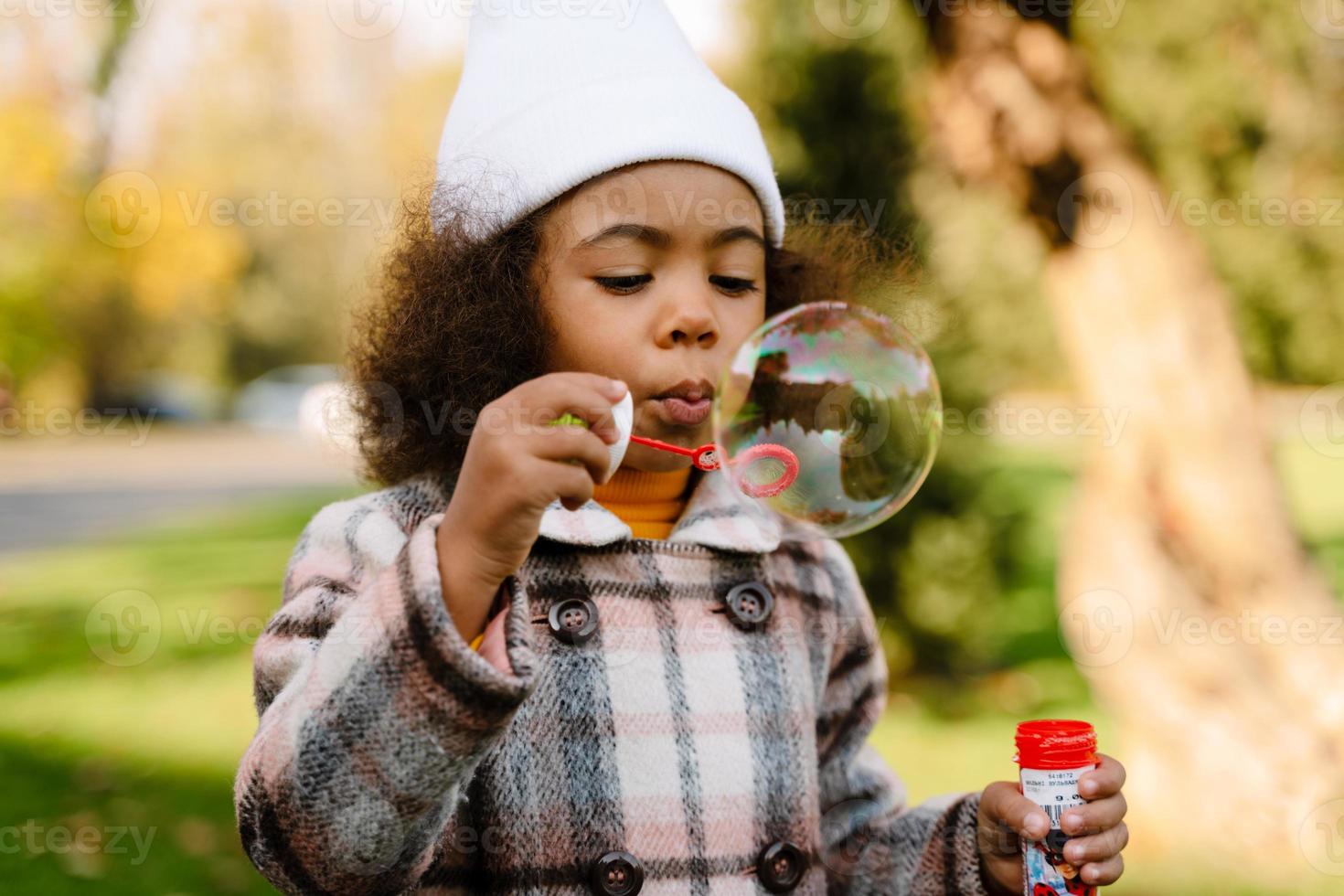 Black girl blowing soap bubbles during walk in autumn park photo