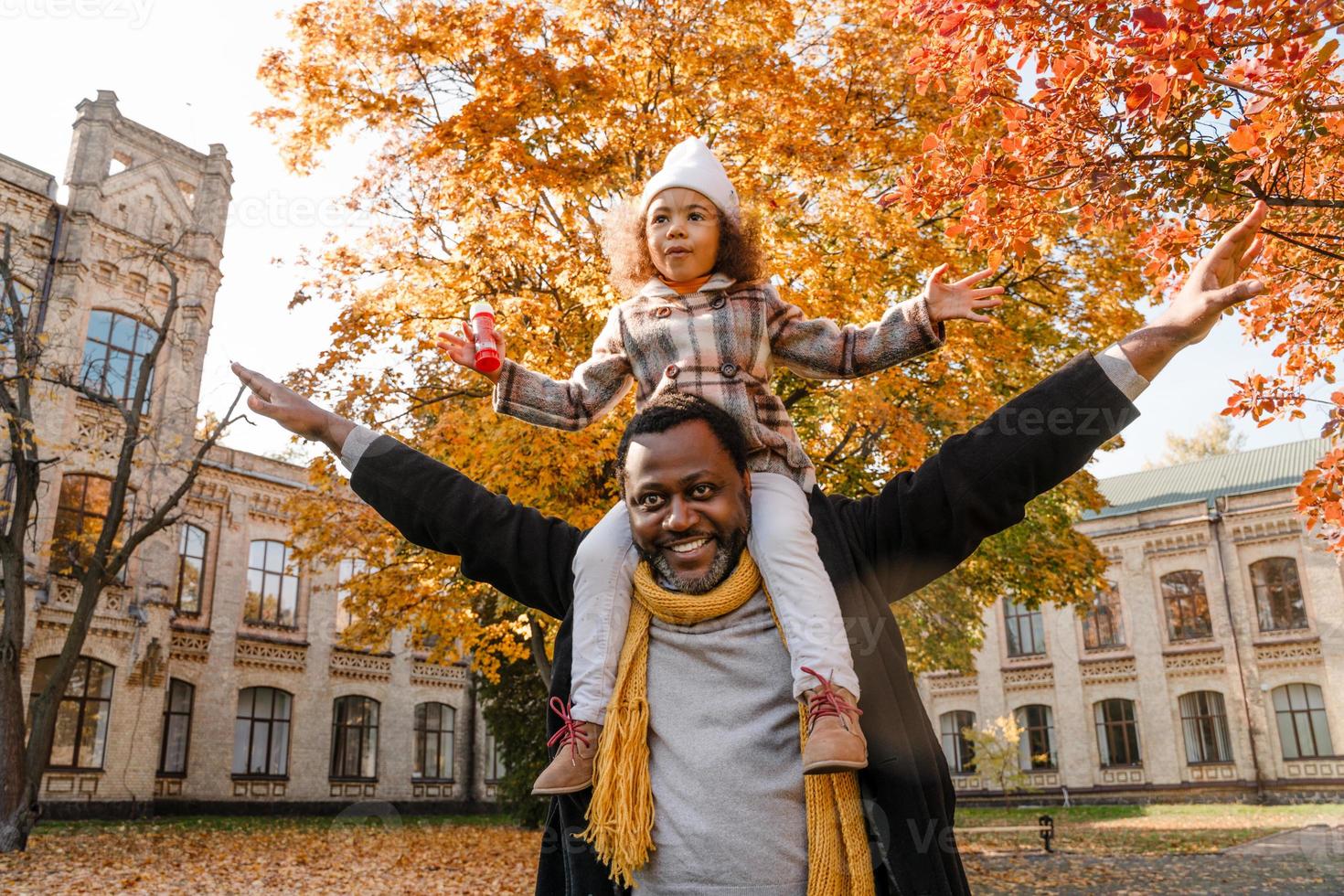 Chica negra divirtiéndose y sentada en el cuello de su abuelo en el parque de otoño foto