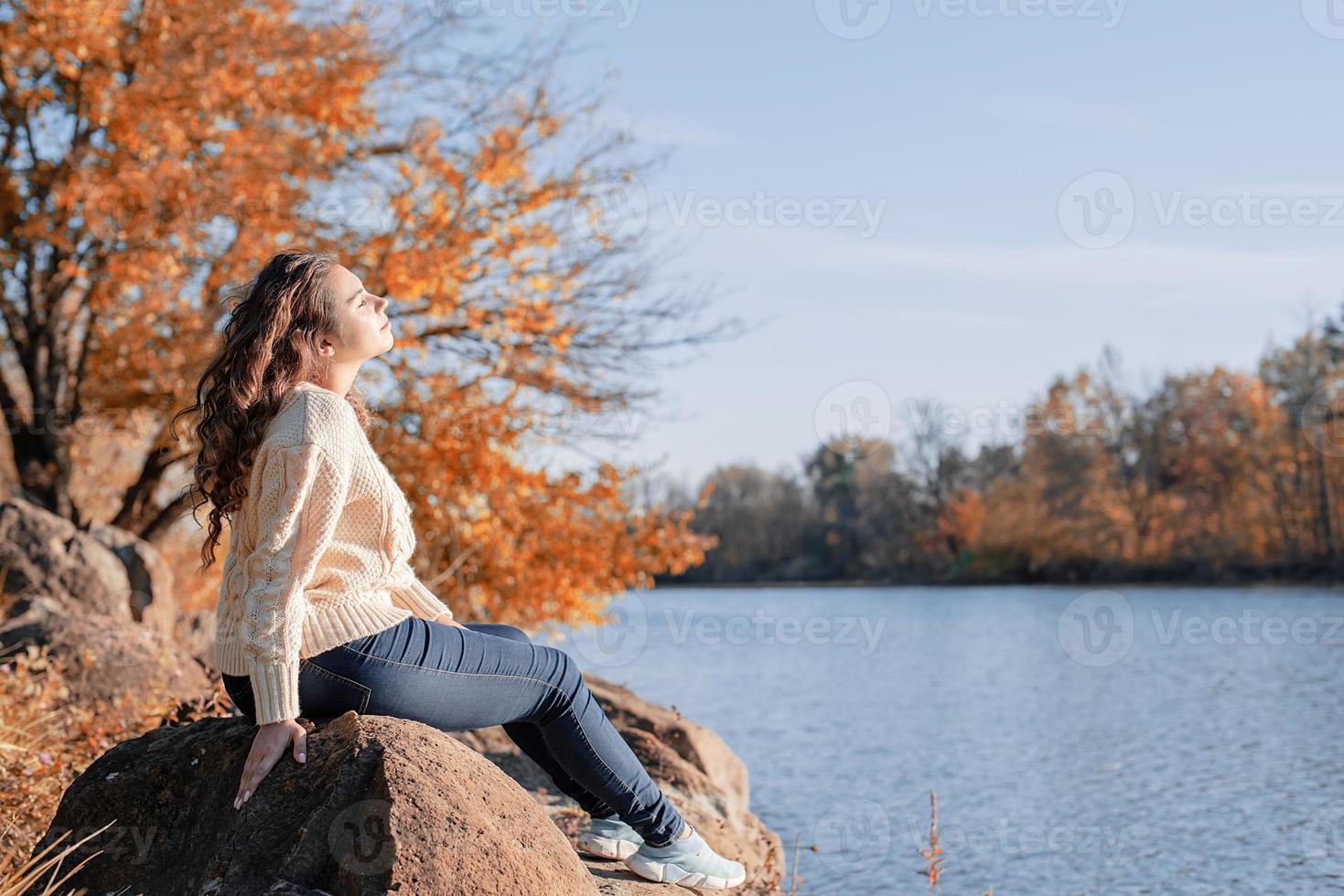 Mujer romántica pensativa sentada sobre las rocas en la orilla del río al atardecer en el día de otoño foto