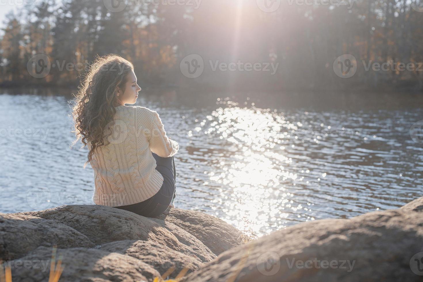 Mujer romántica pensativa sentada en la orilla del río al atardecer en día de otoño foto