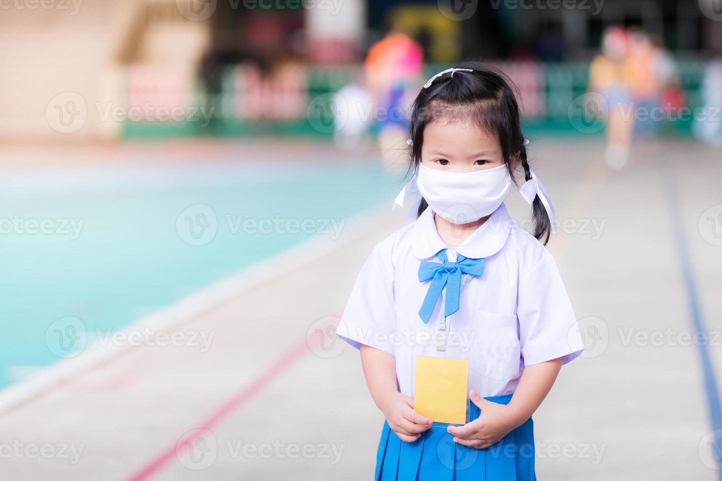 Child wearing white cloth face mask in school uniform is standing. Children go to school with new way of life New Normal. Cute kid looks at the camera. Kindergarten toddler age 3 years old. photo