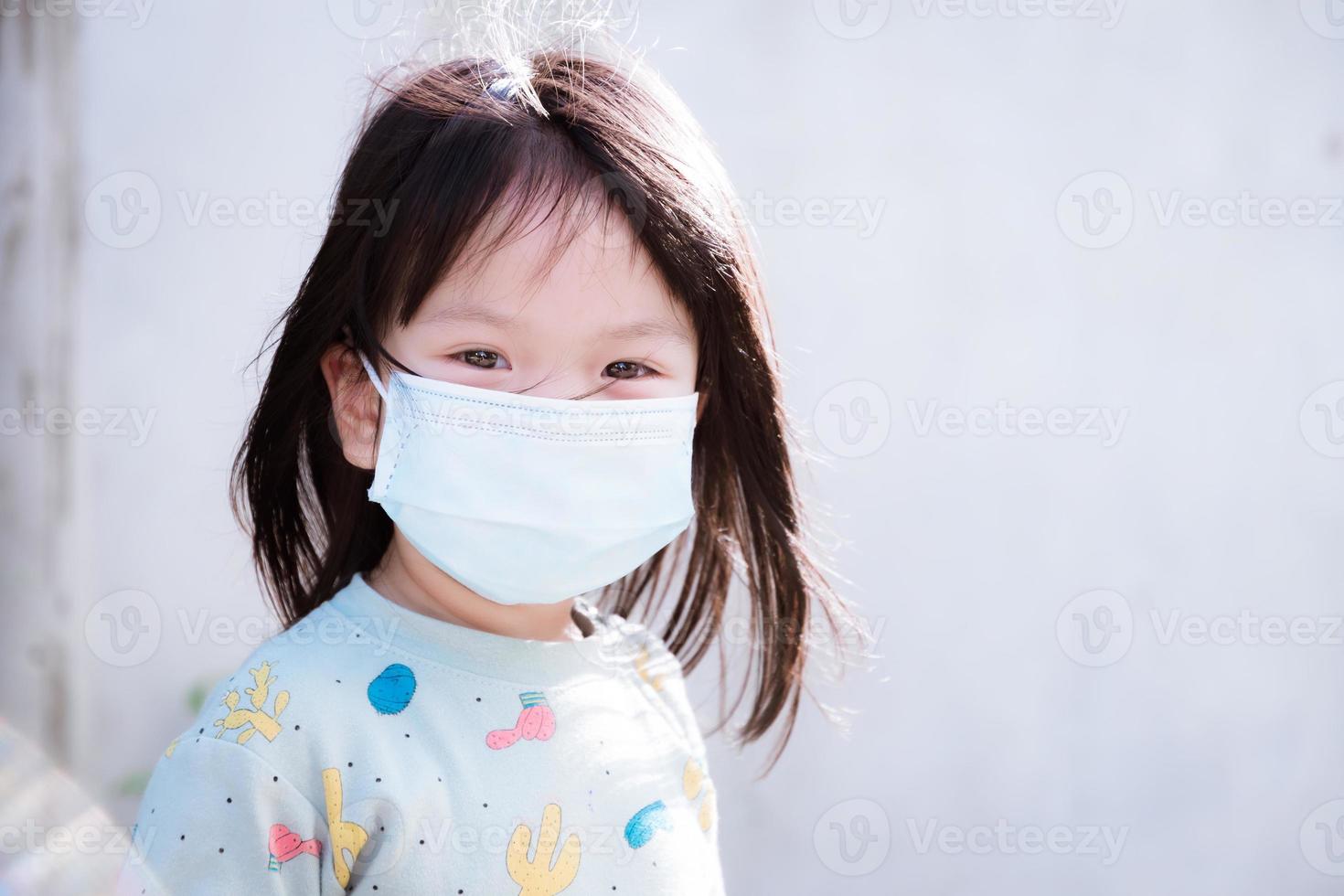 Candid children wear medical face mask to prevent the spread of the coronavirus disease Covid 19. Child wearing pajamas for morning walk in  residential condo area. Cute girl looking at the camera. photo